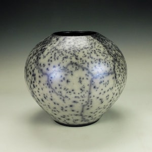 May include: A round ceramic vase with a speckled pattern of black dots on a cream-colored background. The vase has a dark rim and a rounded shape, suitable for home decor.