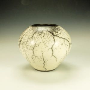 May include: A round, off-white ceramic vase with a dark rim and base. The vase is decorated with a network of black, lightning-like lines and speckled patterns, creating a unique, textured appearance. The vase is a decorative item.