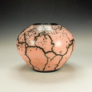 May include: A round, pink ceramic vase with a black crackle pattern. The vase has a dark rim and base, and the surface is speckled with black dots. The overall shape is bulbous, with a slightly tapered base.