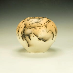 May include: A round, off-white ceramic vase with a unique decorative pattern. The vase features a light tan and black lightning-like design across its surface. The vase has a wide body and a slightly narrower opening at the top.