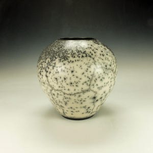 May include: A ceramic vase with a speckled pattern. The vase is primarily off-white with black speckles and a dark rim. The design features a network of fine, dark lines across the surface. The vase has a rounded shape and sits on a dark base.
