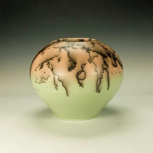 May include: A round, light green ceramic vase with a black and brown crackle glaze. The vase has a light pink hue at the top.