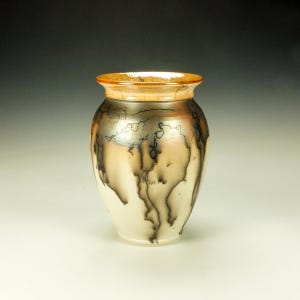 May include: A ceramic vase with a rounded body and a flared rim. The surface features a smooth cream-colored base with dark, wispy black smoke-like patterns and a metallic bronze-toned band around the upper neck and rim area.