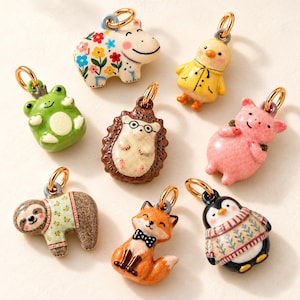 May include: A collection of eight small animal-shaped charms, including a floral hippo, a yellow chick, a green frog, a hedgehog with glasses, a pink pig, a sloth in a sweater, a fox with a bow tie, and a penguin in a patterned sweater.