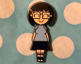 Könnte beinhalten: Eine Emaille-Anstecknadel mit einem Cartoon-Charakter mit schwarzen Haaren, einer Brille, einem blauen Shirt, einem dunkelblauen Rock und rot-weiß gestreiften Socken. Die Anstecknadel befindet sich auf einem blau-weiß gepunkteten Hintergrund.