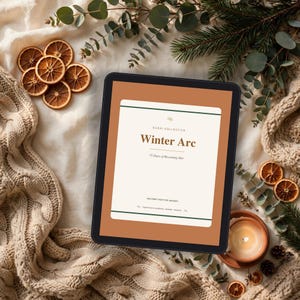 May include: A digital tablet displaying a document titled Winter Arc, 75 Days of Becoming Her by Kudai Collective. The screen features a cream background with dark green borders, surrounded by dried orange slices and a beige knit blanket.