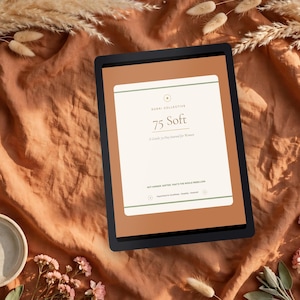 May include: A digital tablet displaying a journal cover titled 75 Soft by Eudai Collective. The screen features a cream-colored rectangle with green horizontal lines, text, and small circular icons against a solid terracotta background.