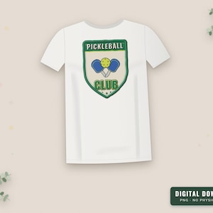 Op de afbeelding: Een wit T-shirt met korte mouwen en een groen, schildvormig embleem op de borst. Het embleem bevat de tekst PICKLEBALL CLUB in groene letters, met twee blauwe rackets en een gele bal in het midden van het ontwerp.