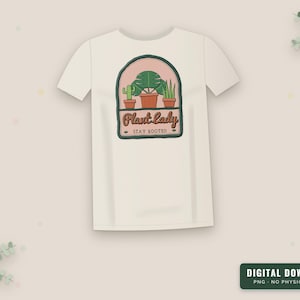 Op de afbeelding: Een crèmekleurig T-shirt met korte mouwen en een afbeelding van drie potplanten in een boogvormig kader. De tekst "Plant lady" en "STAY ROOTED" staat onder de planten in een donkerbruin handschrift en blokletters.