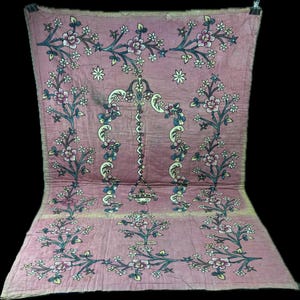 May include: A rectangular pink textile featuring a central arch motif with a hanging lamp design, surrounded by intricate floral patterns in shades of blue, white, and green. The fabric displays a quilted texture with visible stitching throughout.