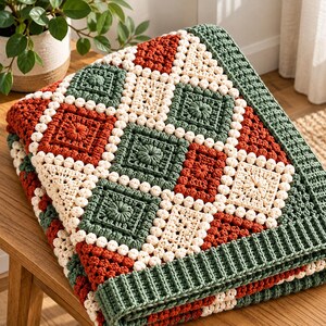 Könnte beinhalten: Eine Häkeldecke mit einem wiederkehrenden Muster aus quadratischen Motiven in Dunkelgrün, Rostorange und Creme. Das Design umfasst strukturierte Noppenmaschen und einen breiten, gerippten dunkelgrünen Rand, der das Stück sauber einfasst.