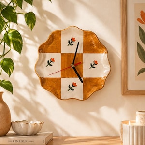 Könnte beinhalten: Eine wellenförmige Wanduhr aus Keramik mit einem Schachbrettmuster aus senfgelben und weißen Quadraten. Jedes weiße Quadrat ist mit einer kleinen, handgemalten orangefarbenen Blume mit grünen Blättern verziert.