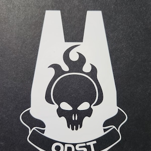 May include: A white vinyl decal featuring a stylized skull with flames rising from the top, set against a dark background. Below the skull is a banner with the text ODST in bold white capital letters, all contained within a shield shape.
