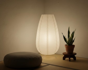 May include: A white paper lantern with a ribbed texture and a teardrop shape stands on thin legs. Beside it, a green snake plant sits in a terracotta pot on a small wooden stand, and a round grey floor cushion rests on the floor nearby.