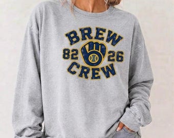 May include: A heather gray long-sleeve sweatshirt featuring a navy blue and yellow graphic on the chest. The design includes the text BREW CREW with the numbers 82 and 26, centered around a stylized baseball glove icon with a baseball inside.