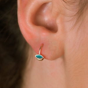 May include: A close-up of a person's ear with a small gold hoop earring featuring a turquoise gemstone. The earring is a simple and elegant design.