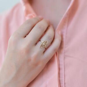 Puede incluir: Un anillo de oro con un dise&ntilde;o delicado e intrincado en forma de &oacute;valo. El anillo se lleva en el dedo de una persona que lleva una camisa rosa.