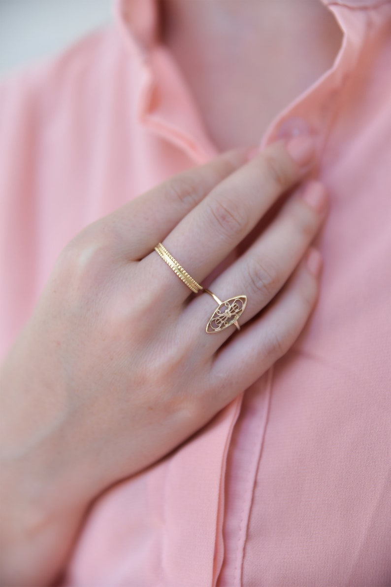 Puede incluir: Primer plano de una mano con dos anillos de oro. El anillo en el dedo &iacute;ndice es una banda delicada, mientras que el anillo en el dedo medio es un dise&ntilde;o m&aacute;s ornamentado con un patr&oacute;n de filigrana.