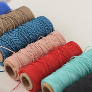 May include: A selection of colourful twine spools in shades of blue, brown, teal, pink, red, and black. The spools are arranged diagonally, displaying the variety of colours and textures. Suitable for crafting and DIY projects.