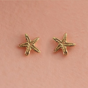 May include: A pair of gold-coloured starfish earrings. The earrings have a textured finish and are displayed on a pale pink surface. These small, delicate earrings would be a great addition to a beach-themed jewellery collection.