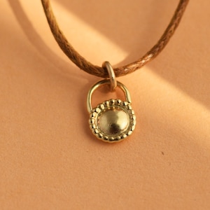 May include: A gold pendant necklace with a round, textured charm. The charm has a small loop at the top and a smooth, circular surface. The necklace is made of brown cord.