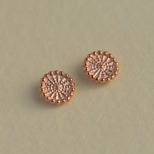 May include: Two small, round, copper-colored metal embellishments with a textured surface. The embellishments have a raised, circular design in the center.