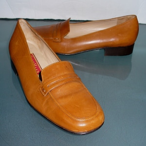 Puede incluir: Un par de mocasines de cuero color canela con tacón bajo. Los zapatos tienen un diseño clásico de mocasín con una correa en el empeine. El interior del zapato es de color claro y la suela es marrón oscuro.