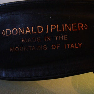 May include: A black leather shoe insole featuring the copper-coloured text DONALD J PLINER, MADE IN THE MOUNTAINS OF ITALY, with a diamond-shaped symbol on each side of the brand name, set against a smooth, dark, and matte-textured background.