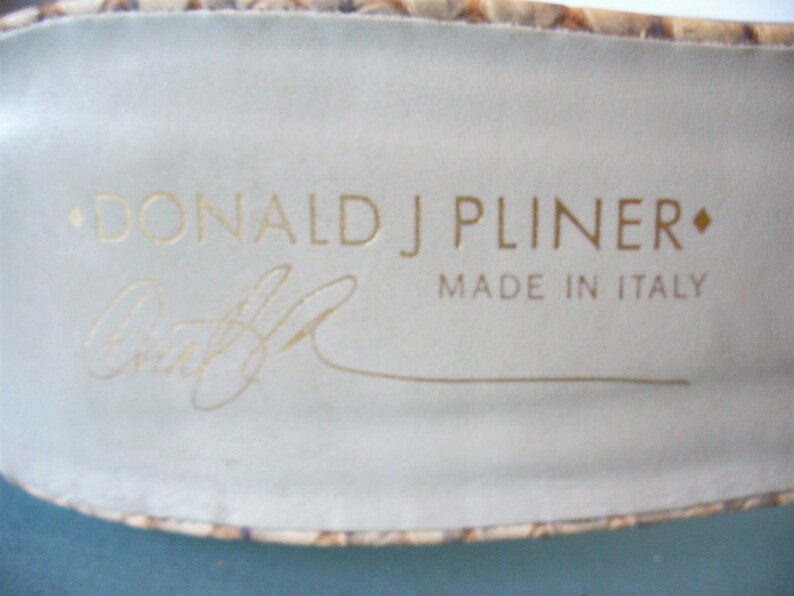 Puede incluir: Primer plano del interior de un zapato con el nombre de la marca "DONALD J PLINER" en letras doradas. Debajo, una firma y las palabras "MADE IN ITALY". El interior del zapato es de color beige claro.