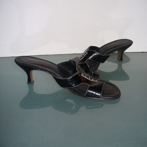 May include: A pair of black patent leather mules with an open toe, a low kitten heel, and a wide strap across the top featuring decorative stitching. The interior footbed is black with gold lettering, and the soles have a smooth, polished finish.