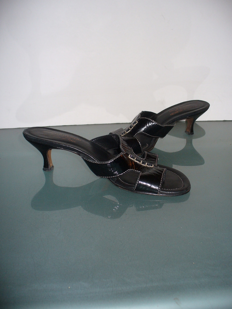 May include: A pair of black patent leather mules with an open toe, a low kitten heel, and a wide strap across the top featuring decorative stitching. The interior footbed is black with gold lettering, and the soles have a smooth, polished finish.
