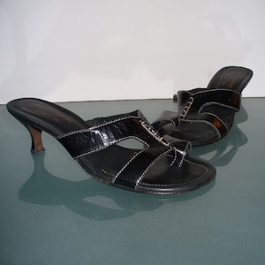 May include: A pair of black leather slide sandals with a toe loop, featuring a kitten heel and white contrast stitching. The sandals have a textured finish on the straps and a branded insole with the text Donald J Pliner Made in Italy.