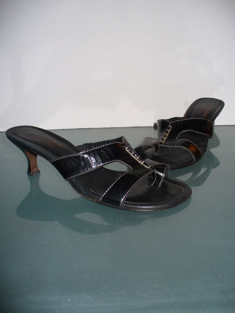 May include: A pair of black leather slide sandals with a toe loop, featuring a kitten heel and white contrast stitching. The sandals have a textured finish on the straps and a branded insole with the text Donald J Pliner Made in Italy.
