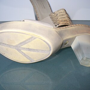 Puede incluir: Sandalia de plataforma con tac&oacute;n transparente. El zapato tiene una parte superior beige claro y una correa con estampado de piel de serpiente. La suela es blanquecina con el nombre de la marca "DONALD J PLINER" impreso. El tac&oacute;n mide unos 7.6 cm.