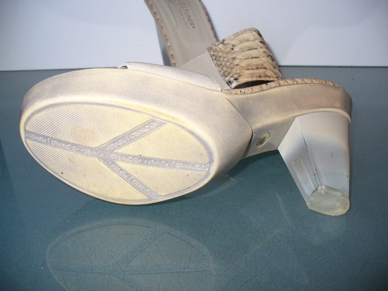 Puede incluir: Sandalia de plataforma con tac&oacute;n transparente. El zapato tiene una parte superior beige claro y una correa con estampado de piel de serpiente. La suela es blanquecina con el nombre de la marca "DONALD J PLINER" impreso. El tac&oacute;n mide unos 7.6 cm.