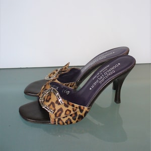 Puede incluir: Un par de sandalias de tac&oacute;n Donald Pliner Couture. Los zapatos de punta abierta presentan una suela de cuero marr&oacute;n, una correa con estampado de leopardo con hebilla plateada y una plantilla morada oscura. Los tacones son negros.