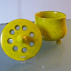 May include: A bright yellow ceramic container with a matching lid. The lid has several circular holes and a central knob. The container has a rounded body and three small feet. The items are displayed on a reflective surface.