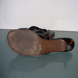 May include: A black sandal with a dark brown sole and a low heel. The upper strap features a glossy, dark tortoiseshell-like finish with visible white stitching along the edges. The sole shows signs of wear with a textured, darkened surface.