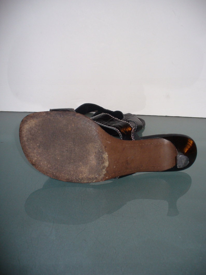 May include: A black sandal with a dark brown sole and a low heel. The upper strap features a glossy, dark tortoiseshell-like finish with visible white stitching along the edges. The sole shows signs of wear with a textured, darkened surface.