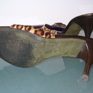 Puede incluir: Un par de mulas de tac&oacute;n alto marrones con la parte superior con estampado de leopardo. Los tacones son de color marr&oacute;n oscuro y curvados. Las suelas son verdes y muestran desgaste. Los zapatos est&aacute;n en &aacute;ngulo.