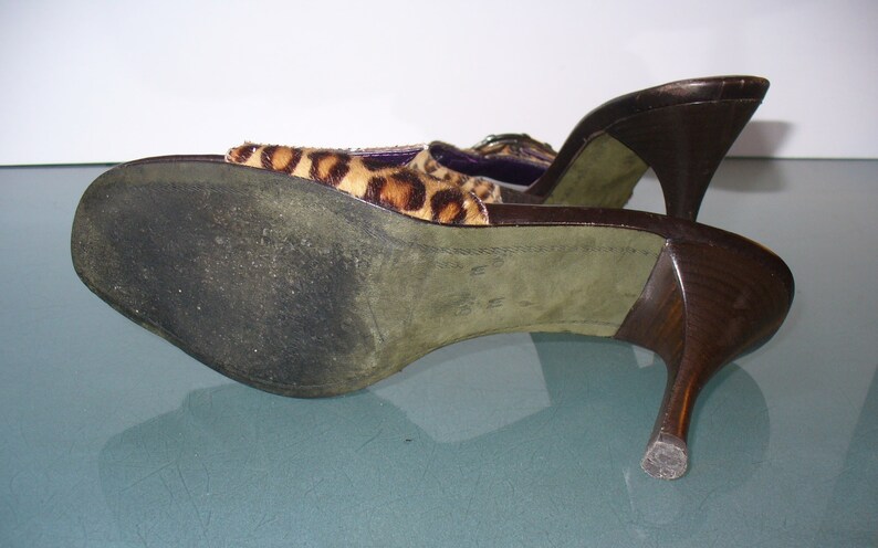 Puede incluir: Un par de mulas de tac&oacute;n alto marrones con la parte superior con estampado de leopardo. Los tacones son de color marr&oacute;n oscuro y curvados. Las suelas son verdes y muestran desgaste. Los zapatos est&aacute;n en &aacute;ngulo.