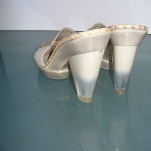 Puede incluir: Un par de mules de tac&oacute;n alto con punta abierta. Los zapatos presentan un tac&oacute;n transparente en forma de cono y una parte superior de color claro con un estampado de piel de serpiente. La marca "GIANNA" es visible en el lateral del zapato.