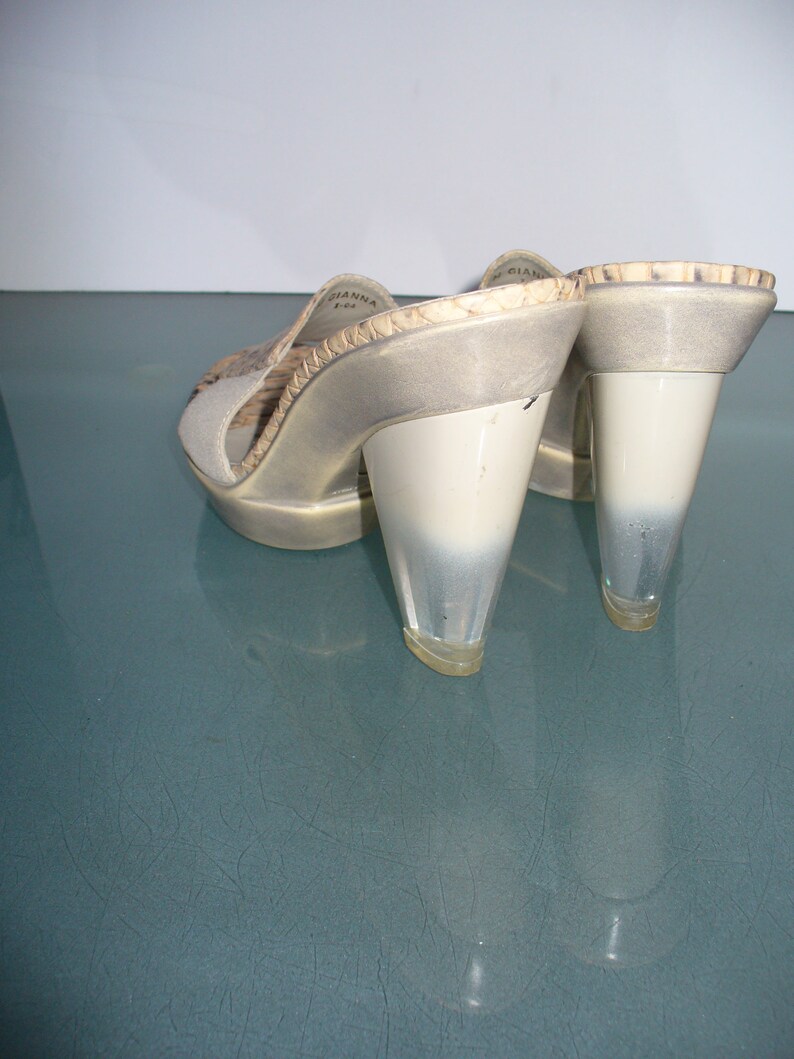 Puede incluir: Un par de mules de tac&oacute;n alto con punta abierta. Los zapatos presentan un tac&oacute;n transparente en forma de cono y una parte superior de color claro con un estampado de piel de serpiente. La marca "GIANNA" es visible en el lateral del zapato.