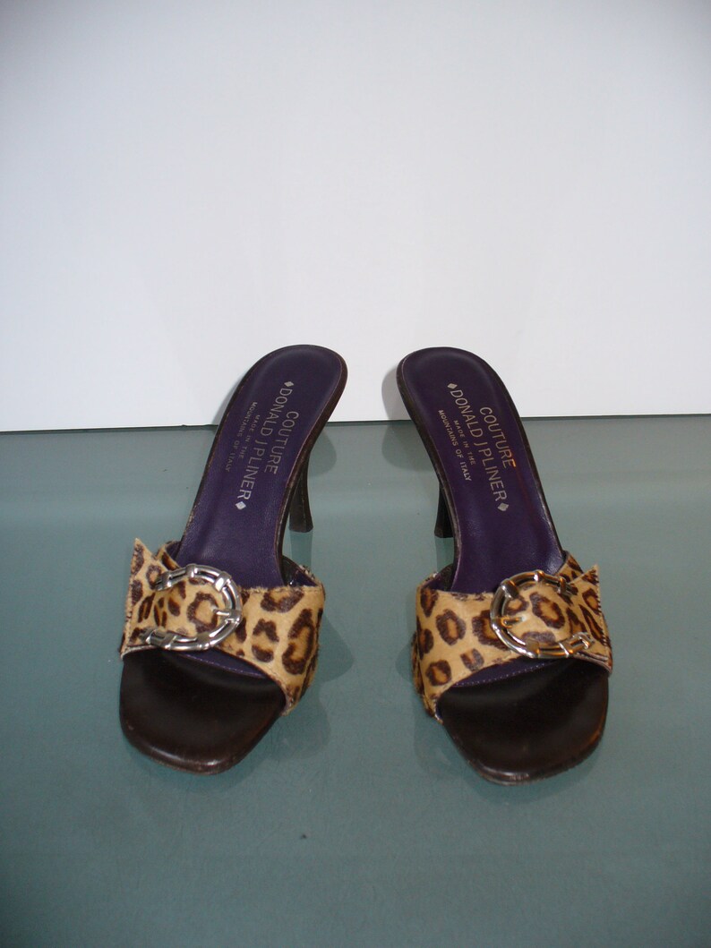 Puede incluir: Un par de sandalias de tac&oacute;n Donald J Pliner Couture. Los zapatos de punta abierta presentan una suela de cuero marr&oacute;n, una correa con estampado de leopardo con hebilla plateada y una plantilla morada oscura. Los tacones miden unos 10 cm.