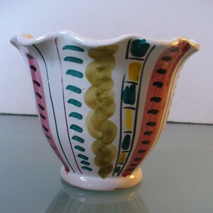 May include: A white ceramic vase with a scalloped rim and a flared shape. The vase is decorated with vertical stripes of pink, teal, and orange, along with yellow and green swirled and rectangular patterns. The base is a small, rounded foot.