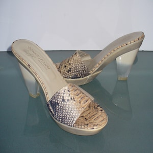 Puede incluir: Sandalias sin tal&oacute;n con estampado de piel de serpiente en las correas. Las sandalias tienen tacones transparentes en forma de cu&ntilde;a y una plantilla beige. La marca "DONALD J PLINER" es visible en la plantilla. Hecho en Italia.