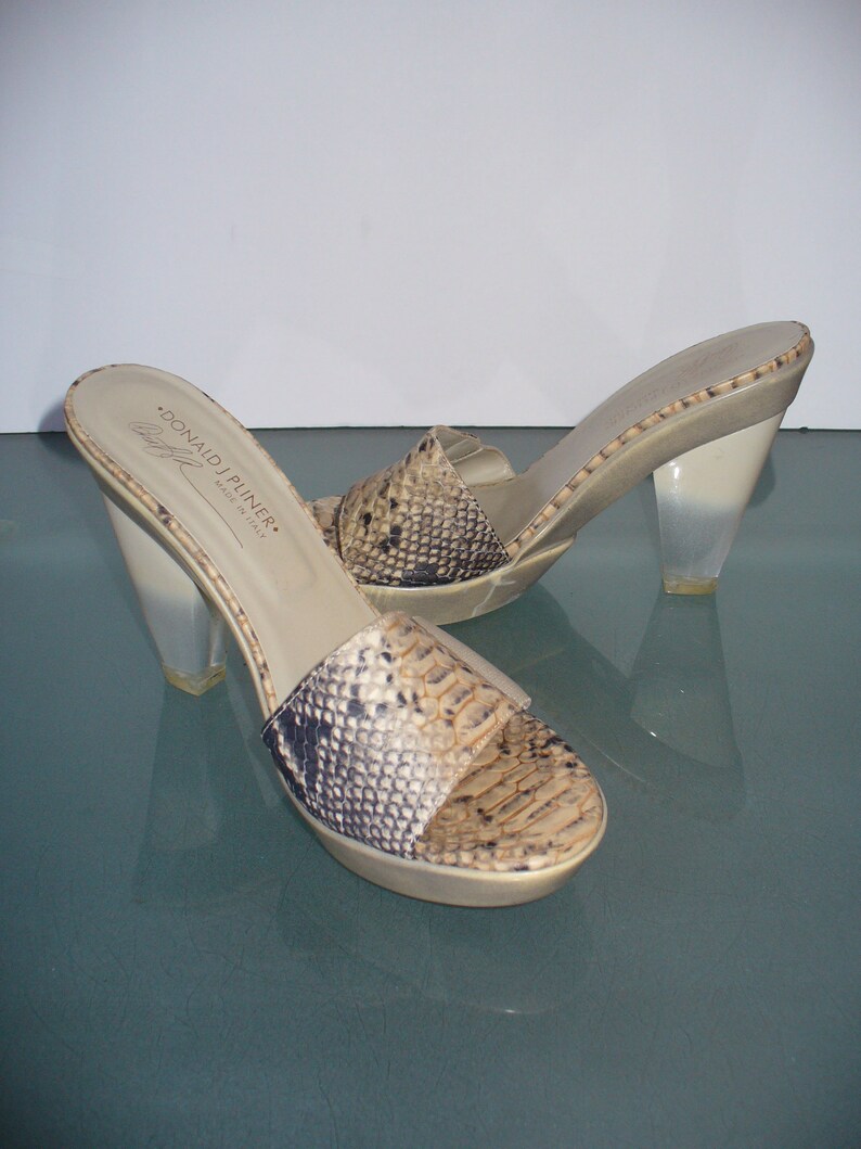 Puede incluir: Sandalias sin tal&oacute;n con estampado de piel de serpiente en las correas. Las sandalias tienen tacones transparentes en forma de cu&ntilde;a y una plantilla beige. La marca "DONALD J PLINER" es visible en la plantilla. Hecho en Italia.