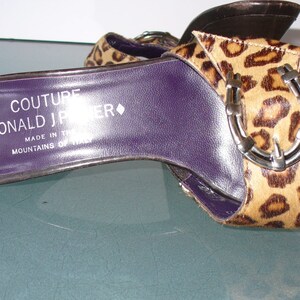 Puede incluir: Sandalias de punta abierta con estampado de leopardo. La plantilla morada muestra el texto "COUTURE DONALD J PLINER MADE IN ITALY MOUNTAINS OF ITALY". Las sandalias tienen una hebilla plateada.