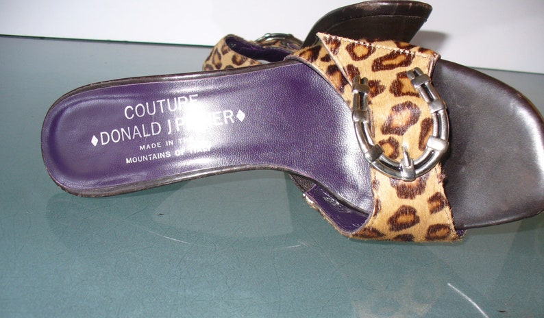 Puede incluir: Sandalias de punta abierta con estampado de leopardo. La plantilla morada muestra el texto "COUTURE DONALD J PLINER MADE IN ITALY MOUNTAINS OF ITALY". Las sandalias tienen una hebilla plateada.