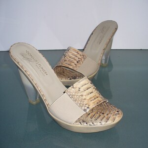 Puede incluir: Un par de mulas Donald J Pliner con punta abierta. Los zapatos presentan un estampado de piel de serpiente en la parte superior, una correa el&aacute;stica beige y tacones de acr&iacute;lico transparente. Las suelas son beige, hechas en Italia.